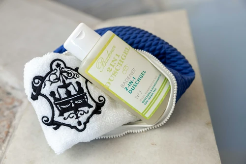 Blaue Badetasche mit hellgrünem Duschgel und weissem Handtuch mit schwarzer Stickerei liegend auf hellem Steinboden