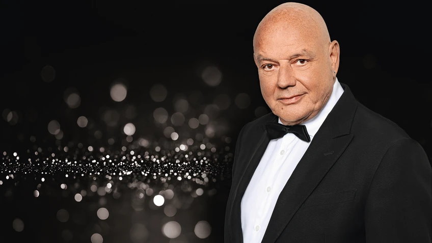 Wolfgang Böck steht elegant gekleidete mit Glatze und schwarzem Smoking vor festlichem Hintergrund und blickt ruhig und selbstbewusst in die Kamera.