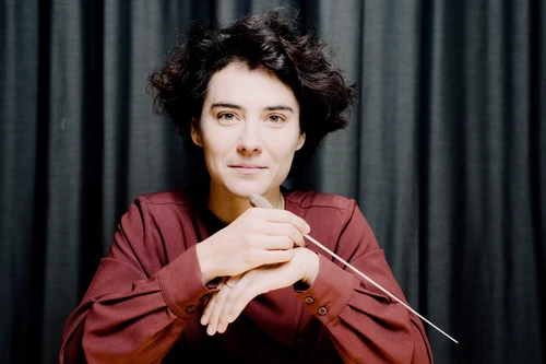 Frau mit lockigen dunklen kurzen Haaren lächelt, in der Hand hält sie einen Dirigentenstock