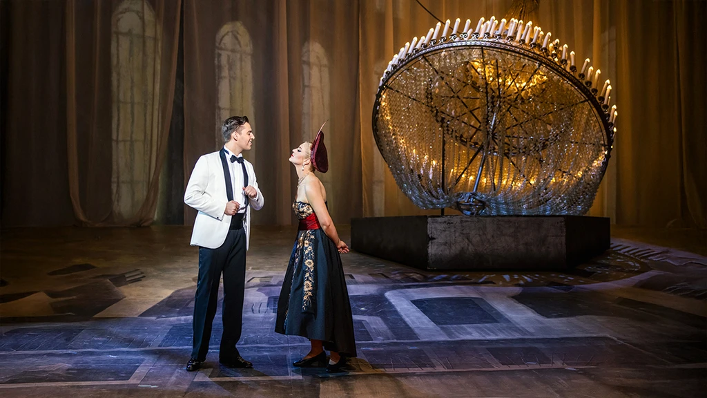 Zwei Darsteller stehen auf einer Bühne vor einem großen, auf dem Boden liegenden kunstvollen Kronleuchter mit vielen Kerzen. Die Person links trägt einen weißen Smoking mit schwarzer Fliege, die Person rechts ein elegantes schwarzes Kleid mit rotem Oberteil und Kopfschmuck. Sie diskutieren miteinander. Der Hintergrund zeigt hohe Fenster und warme Beleuchtung.
