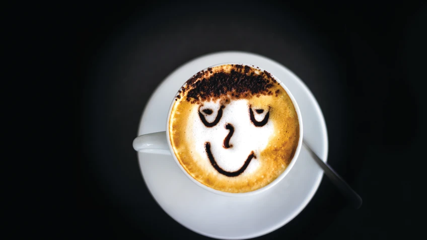 Eine Tasse Cappuccino steht auf dunklem Untergrund, wobei der Milchschaum mit Kakaopulver ein lächelndes Gesicht mit geschlossenen Augen und Haaren bildet.