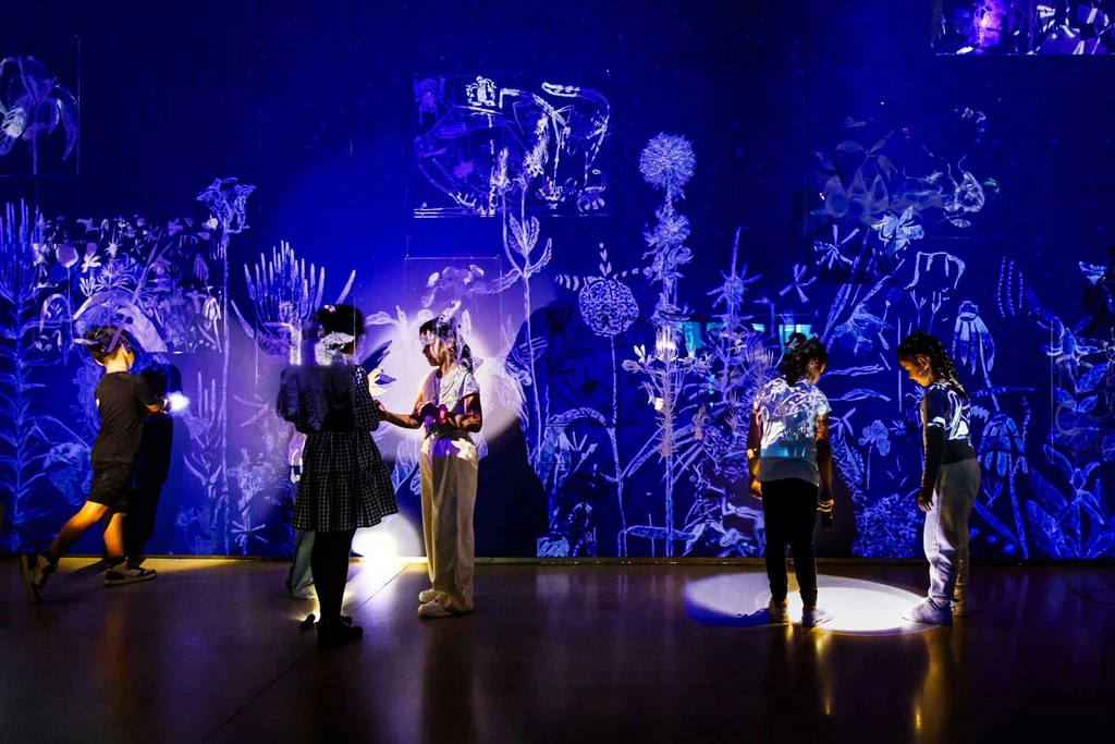 Five children shine torches in a dark room. Behind them is a dark blue wall with white nature projections. In front of them hang acrylic elements with engraved drawings by Robert Gabris, Iva Ivanova and Theodor Moise.