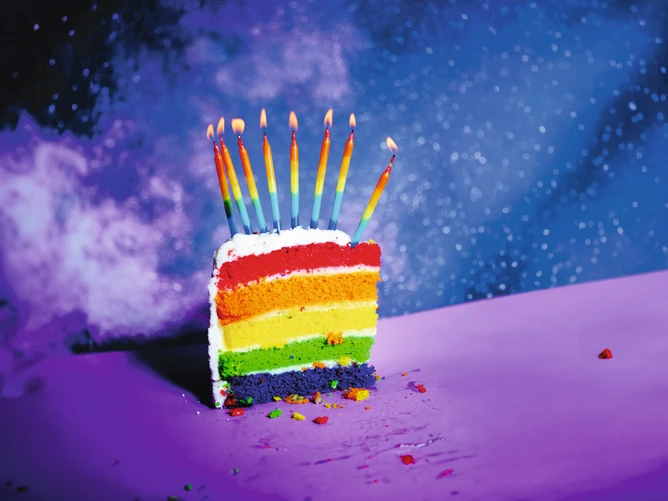 Bild von einem Tortenstück in Regenbogenfarben mit acht bunten brennenden Kerzen darauf