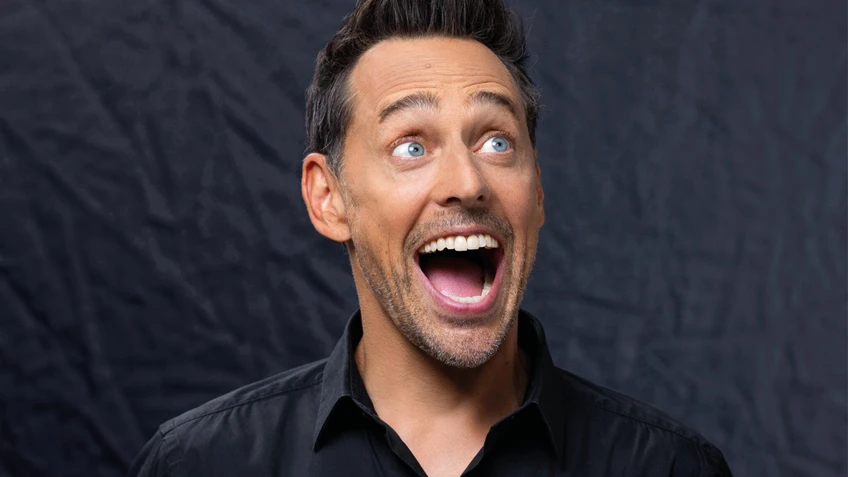 Tricky Niki steht mit schwarzem Hemd vor dunklem Hintergrund und lacht mit weit geöffnetem Mund nach rechts oben, als wäre er überrascht oder begeistert.