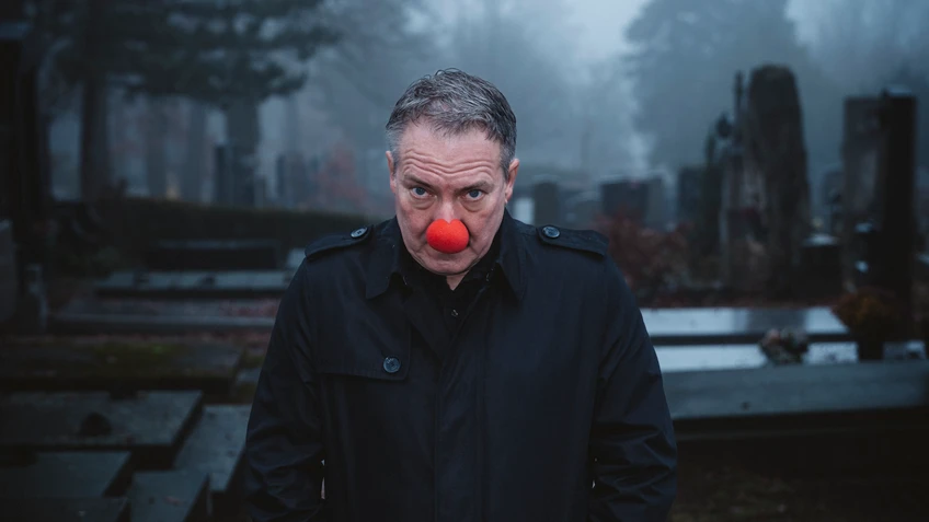 Robert Palfrader steht mit schwarzem Mantel und roter Clownsnase nach vorn gebeugt und ernst blickend auf einem nebligen Friedhofsweg zwischen Gräbern.