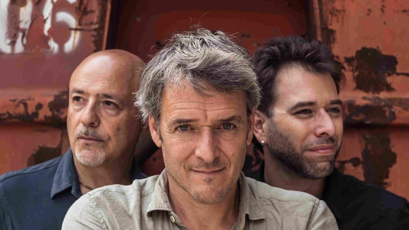 Christof Spörk und die Fratelli Bros. stehen eng beisammen vor einer rostigen Metallwand, wobei Spörk in der Mitte mit verschränkten Armen direkt in die Kamera blickt und die beiden anderen leicht versetzt hinter ihm stehen.