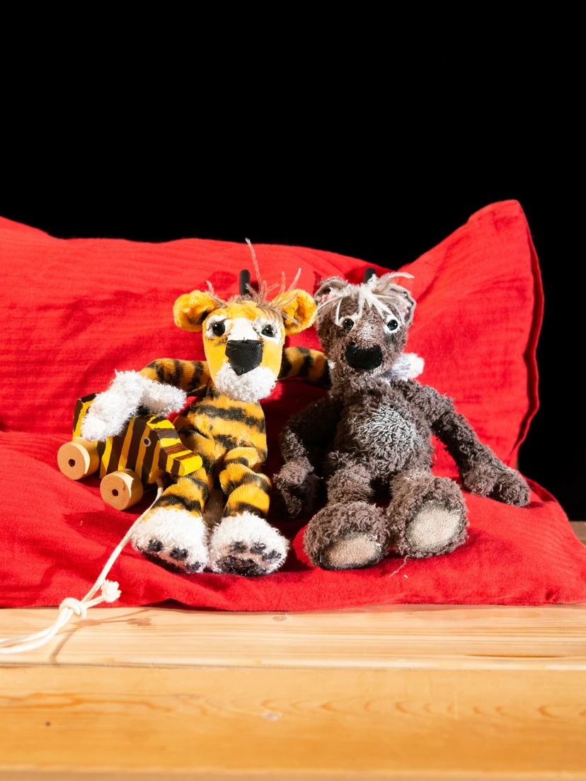 Zwei weiche Tierpuppen – ein Tiger mit Tigerente an der Hand und ein Bär – sitzen eng nebeneinander auf einem roten Kissen auf einem Holztisch vor schwarzem Hintergrund.