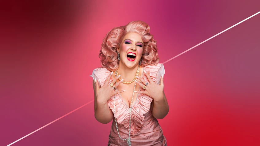 Nahaufname einer Frau in rosa Rüschenkleid, langer Perlenkette, rosa Perücke die vor einem rosafarbenen Hintergrund zur Kamera nach vorne gebeugt steht. Sie lacht mit weitgeöffnetem Mund.