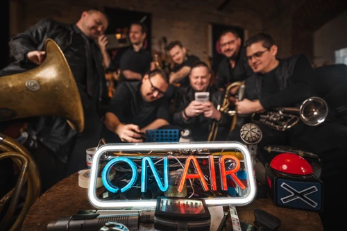 Musiker sitzen rund um ein Leuchtschild mit der Aufschrift "on air", Instrumente und andere Gegenstände unscharf
