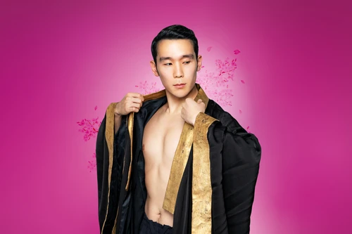 Eine Person in einem schwarzen, goldverzierten Umhang öffnet diesen leicht und blickt zur Seite, vor einem pinkfarbenen Hintergrund mit schwebenden Blütenblättern.