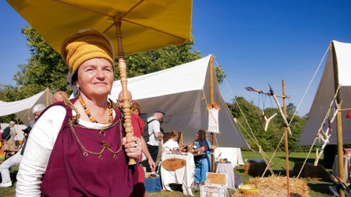 Eine Keltin geht bei Verkaufständen vorbei, sie trägt einen Hut und einen Sonnenschirm 