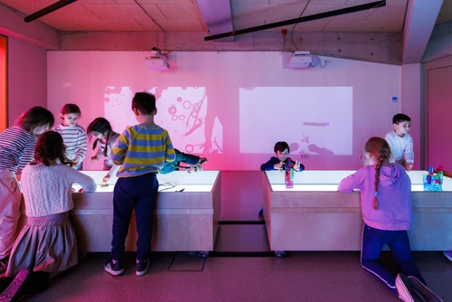 Eine Gruppe von Kindern spielt in einem abgedunkelten Raum an leuchtenden Tischen. Einige bauen mit bunten Bausteinen, andere experimentieren gemeinsam an einem Lichttisch. An der Wand werden farbige Projektionen sichtbar, die von den Aktivitäten der Kinder stammen. Die Atmosphäre wirkt lebendig und spielerisch.