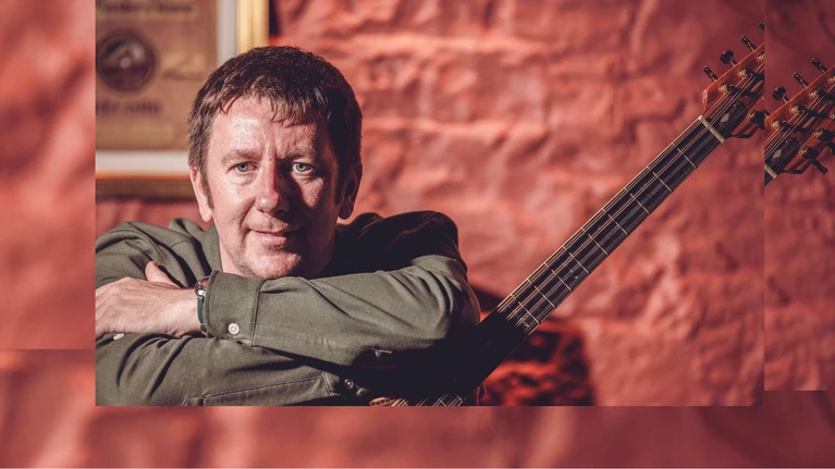 A photograph of the musician Daoirí Farrell with his instrument, a stringed instrument known as a bouzouki, in front of a red wall.