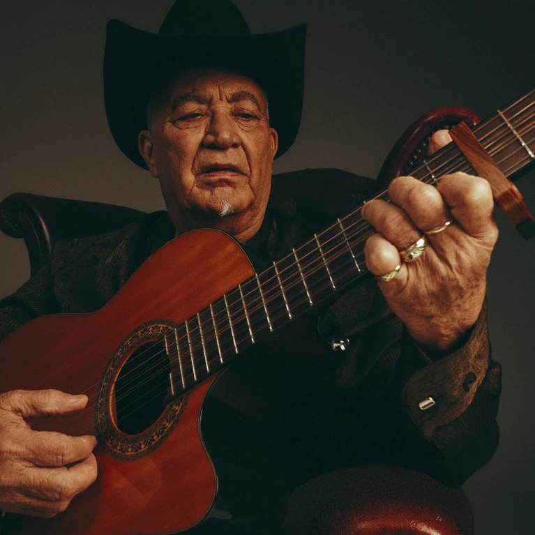 A portrait photograph of musician Elias Ochoa with his instrument, an acoustic guitar, wearing a black suit and a black hat.