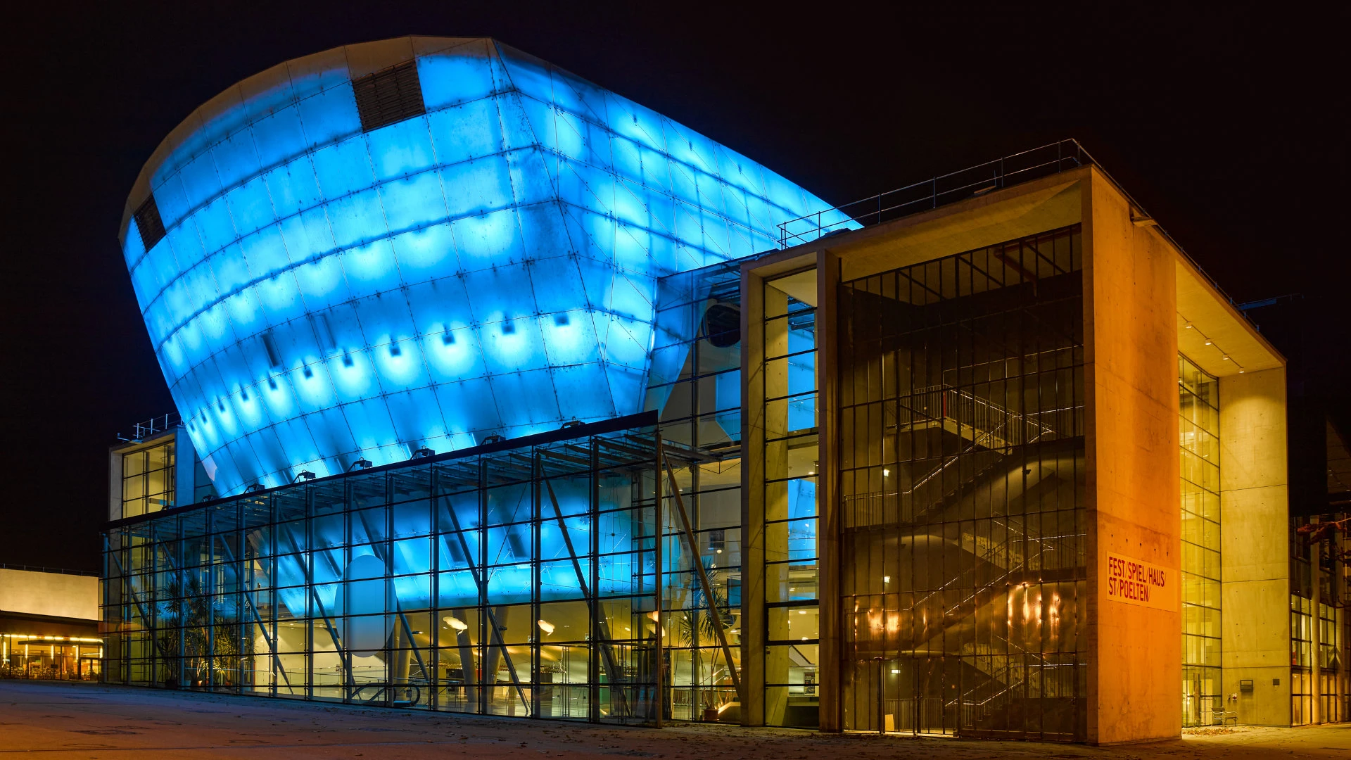 Modernes Festspielhaus mit markanter, blau beleuchteter Fassade und verglastem Eingangsbereich bei Nacht.