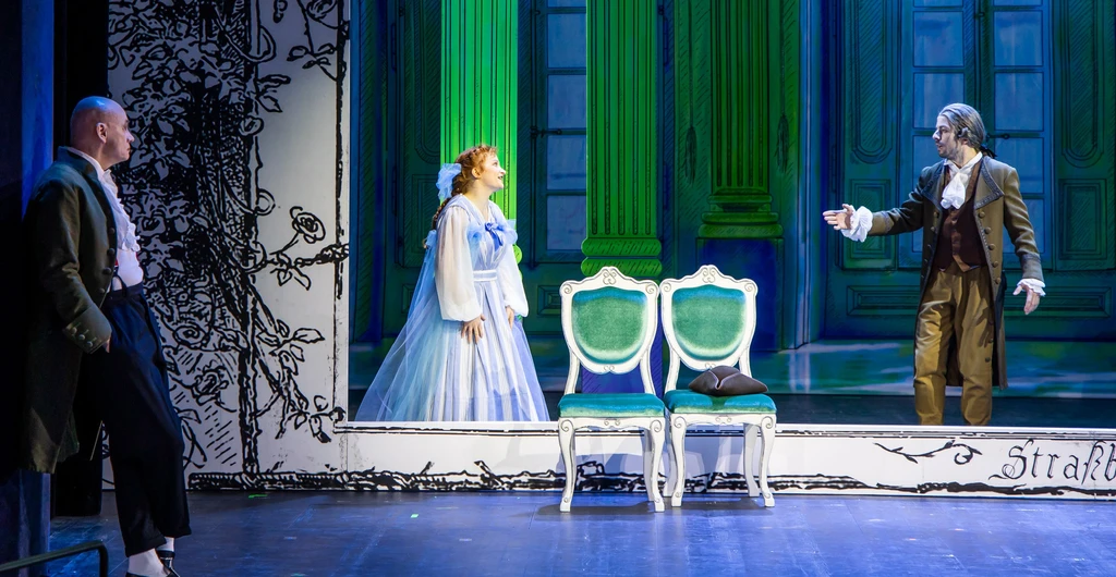 Theaterszene in romantischer Kulisse, weiß-grün. Ein Mann deutet mit einem Arm auf eine Frau in langem hellblauen Kleid. Links im Vordergrund lehnt ein Mann, seitlich sichtbar und beobachtet die Szenerie.