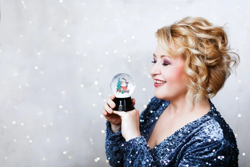 Frau in dunkelblauem Pailletenkleid, blonden Locken und roten Lippen, hält eine Schneekugel