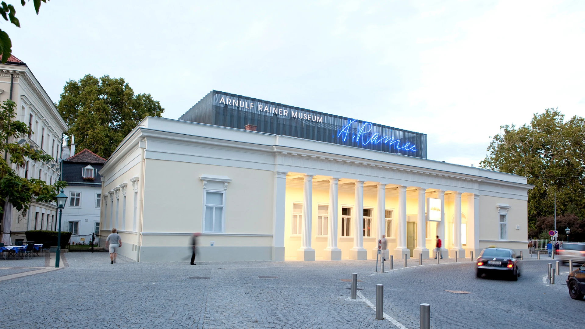 Frontansicht des Arnulf Rainer Museums mit klassizistischer Fassade und beleuchtetem Eingangsbereich, darüber moderne Neonaufschrift.
