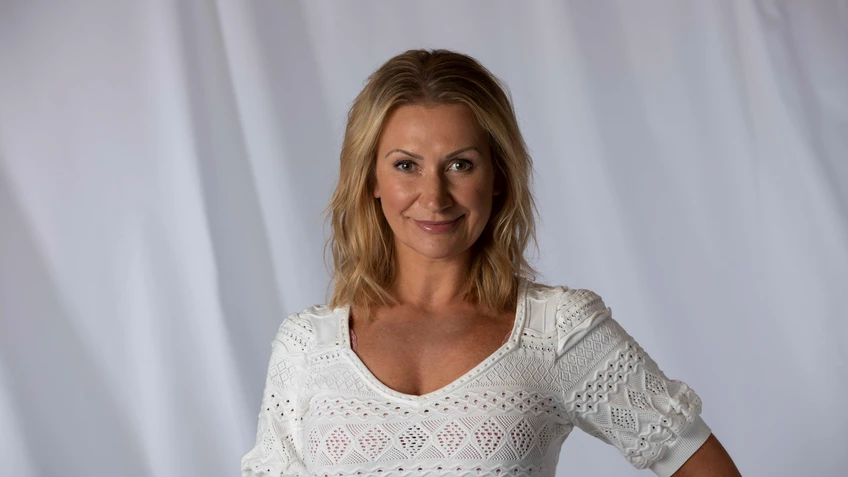 Angelika Niedetzky steht lächelnd mit schulterlangen blonden Haaren in einem weißen, gemusterten Spitzenoberteil vor einem neutralen hellen Hintergrund und blickt charmant in die Kamera.