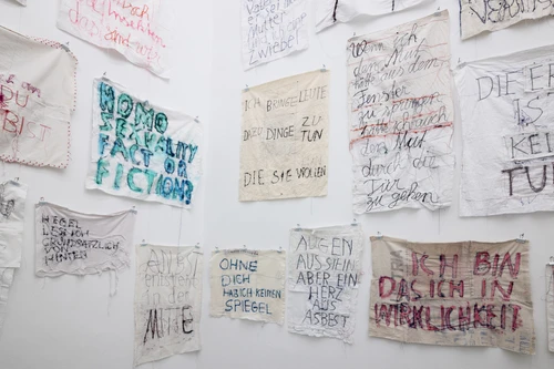 Wandinstallation aus zahlreichen weißen Stoffstücken, auf die handschriftlich Sätze und Wörter in verschiedenen Farben gestickt oder gemalt sind. Die Tücher hängen dicht nebeneinander und zeigen Botschaften in unterschiedlichen Stilen, teilweise farbig hervorgehoben oder mehrfach überarbeitet.