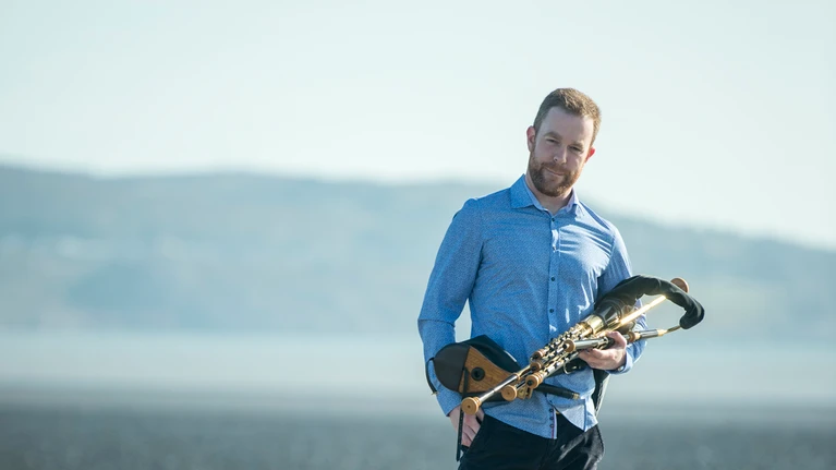 Foto von Musiker Mark Redmond mit seinem Instrument, mit Uilleann Pipes, zu sehen am Strand, hinter ihm das Meer.