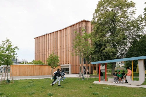 Modernes Gebäude mit vertikaler Holzfassade inmitten einer Grünfläche, Kinder spielen und sitzen im Vordergrund.