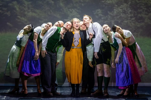 A group of people in colourful, historical clothing, with a young woman in the centre whom the others are facing. They are all standing close together, leaning towards the woman in the middle, creating a strong sense of community and cohesion. The dark stage and the targeted lighting draw attention to the closed group formation and the emotional expression of the scene.