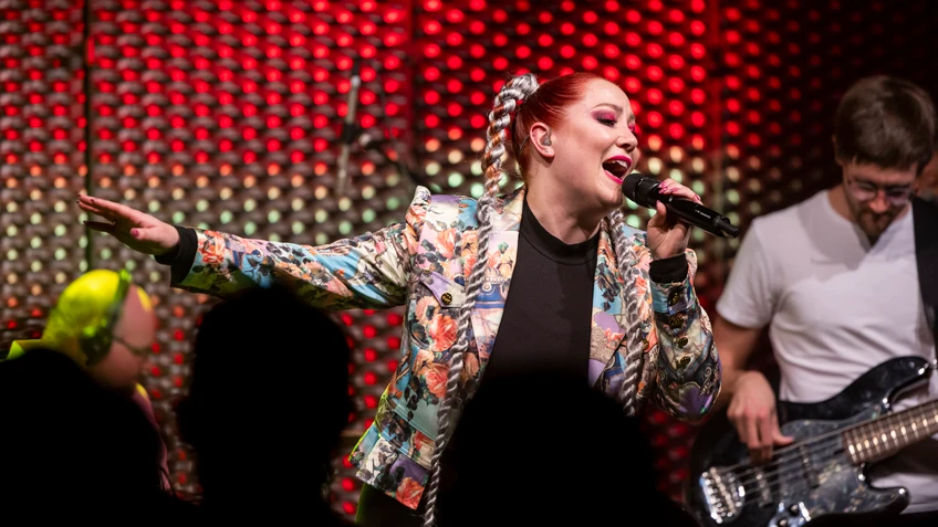 Eine Sängerin mit sehr langen Zöpfen und pinkem Lidschatten singt in ein Mikrophon, ein Gitarrist im Hintergrund