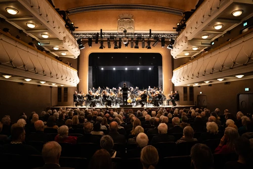 Saal eines Theaters voll mit Publikum, auf der Bühne ein Symphonieorchester im Scheinwerferlicht