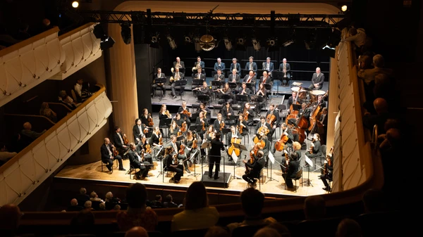 Saal eines Theaters voll mit Publikum, auf der Bühne ein Symphonieorchester im Scheinwerferlicht