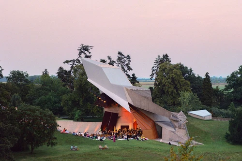 Architektonisch außergewöhnliche Freiluftbühne mit Konzertbesuchern im Grünen bei Sonnenuntergang.