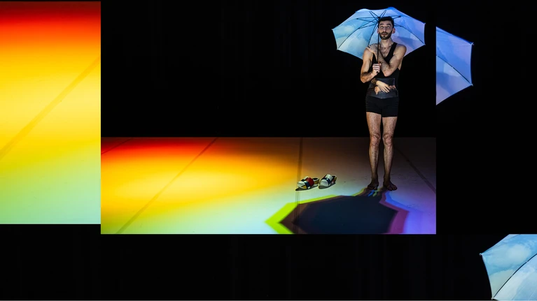 Foto aus der Performance "Souvenirshop" performt von BRONKS & Not Standing, choreografiert von Alexander Vantournhout. Zu sehen ist ein in schwarz gekleideter Mann mit Regenschirm, auf einer fast ganz dunklen Bühne - auf dem Boden bricht sich Licht zu einem Regenbogen.