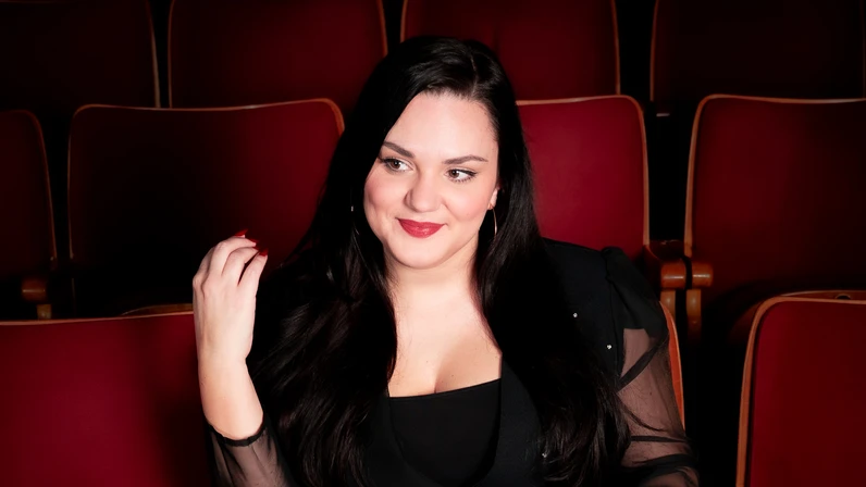 Ina Jovanovis sitzt elegant gekleidet, mit langen dunklen Haaren lächelnd und entspannt in einem leeren Theater zwischen roten Samtsesseln.