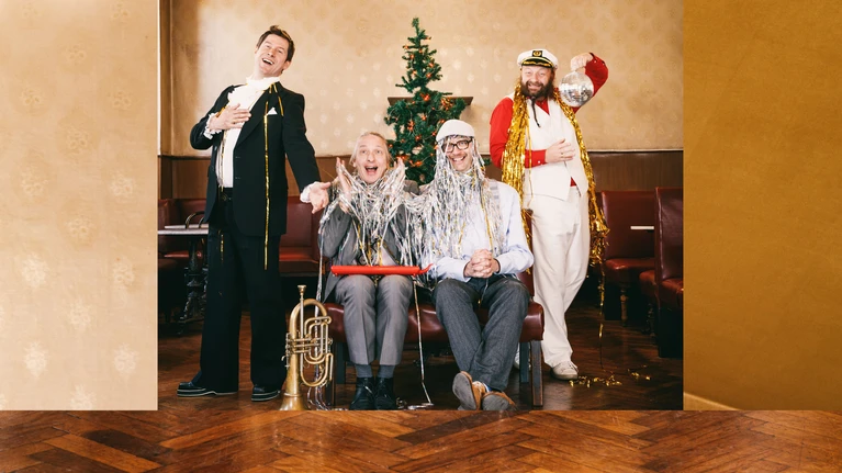 A photo of the musicians Sebastian Fuchsberger, Leonhard Paul, Michael Hornek and Thomas Gansch in an empty coffee house, with a Christmas tree behind them; the cheerful figures are partly draped in tinsel.