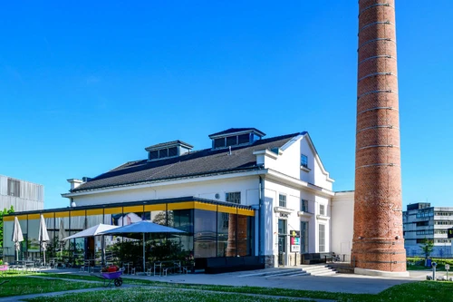 Historisches Industriegebäude mit hohem Ziegelkamin und angeschlossenem Cafébereich mit Sonnenschirmen.
