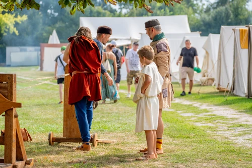 Personen in antikem Gewand stehen um eine Nachbildung einer antiken Maschine. Im Hintergrund sieht man Besucher und Zelte.