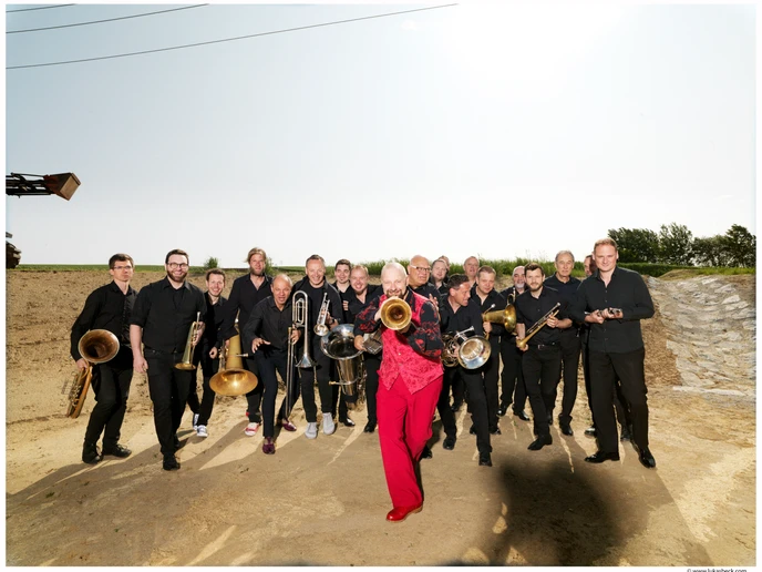 Eine große Gruppe Musiker mit Blasinstrumenten, vorne ein Mann in roter Hose und rotem  Gilet mit Flügelhorn