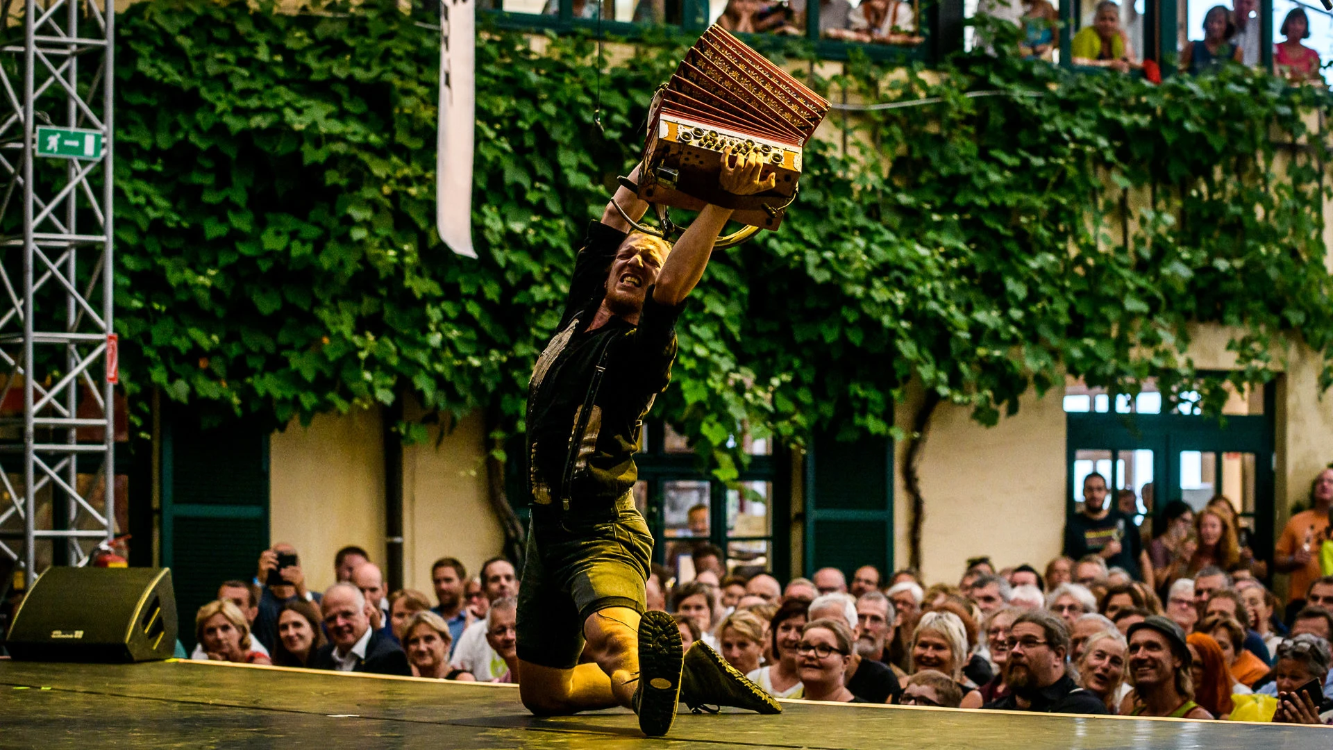 Darbietung eines Musikers mit Akkordeon vor dicht gedrängtem Publikum in grün bewachsenem Innenhof.