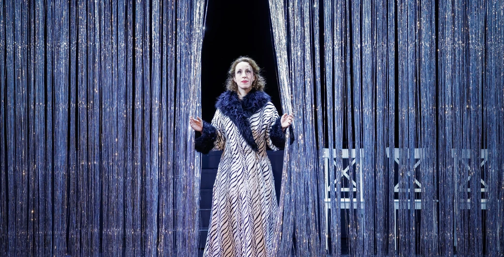 Frau steht in der Mitte eines Glitzervorhangs und hat ihn zur Seite geschoben, sie trägt einen Mantel mit gestreiftem Muster mit schwarzem Pelzkragen