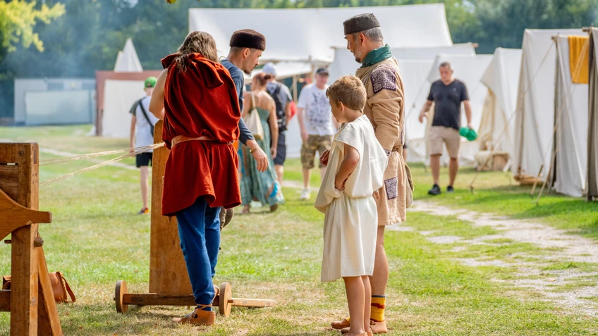 Personen in antikem Gewand stehen um eine Nachbildung einer antiken Maschine. Im Hintergrund sieht man Besucher und Zelte.