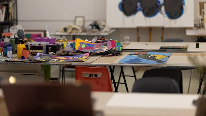 Blick in ein Atelier mit Arbeitstischen, Stühlen und bunten Materialien; im Hintergrund sind Werkzeuge und Arbeitsflächen zu sehen.