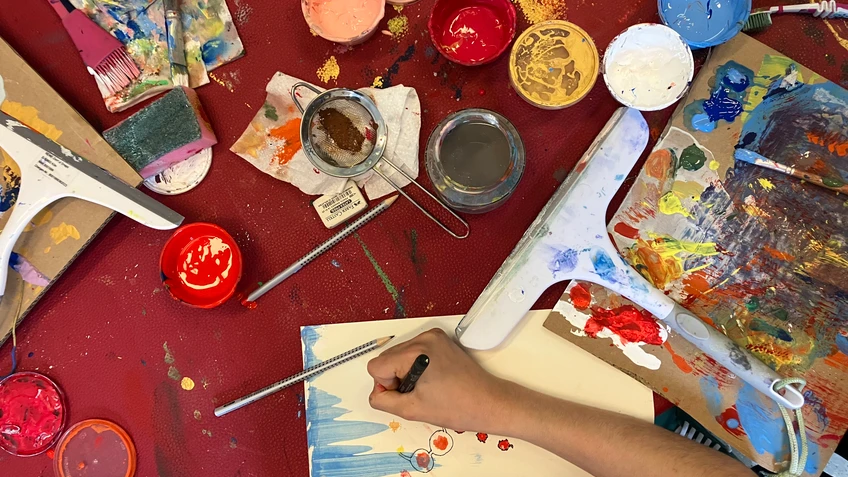 Blick von oben auf eine kreative Arbeitsszene mit Farben, Pinseln und einer Person, die auf Papier malt.