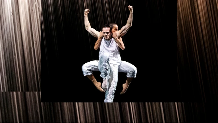 Zwei Tänzer:innen performen "Mémoires d’Oubliettes", eine Choreografie von Jirí Kylián. Ein Tänzer in weißem Kostum steht im Vordergrund auf einem Bein, das andere vor sich angewinkelt, seine Arme seitlich nach oben, die Armmuskeln stark angespannt. Hinter ihm ist eine weitere Person zu sehen, ebenfalls in weißem Kostüm, die sich an seinem Rücken festklammert, beide Beine angehoben und seitlich angewinkelt.