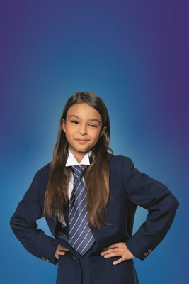 Circa neunjähriges Mädchen mit langen Haaren vor blauem Hintergrund, Hände in die Hüfte gestemmt. Trägt Anzugjacke und blau-weiß gestreifte Krawatte. Nahaufnahme.