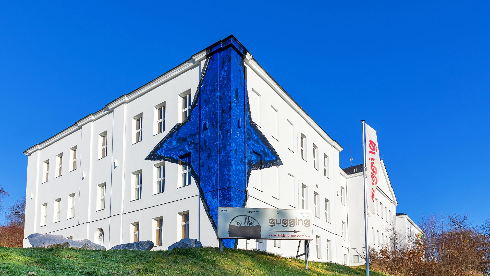 Weißes Museumsgebäude mit auffälligem blauen Wandgemälde in Pfeilform und Beschilderung Gugging.