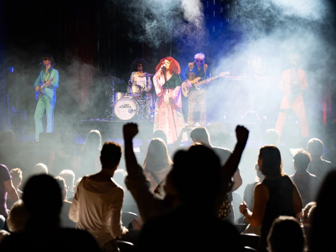 Eine bunt gekleidete Band auf der Bühne, Rauchschwaden, Publikum tanzend im Zuschauerraum