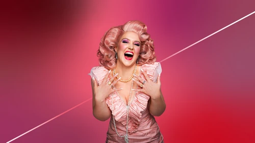 Nahaufname einer Frau in rosa Rüschenkleid, langer Perlenkette, rosa Perücke die vor einem rosafarbenen Hintergrund zur Kamera nach vorne gebeugt steht. Sie lacht mit weitgeöffnetem Mund.