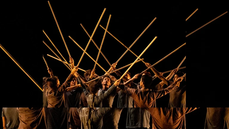 Performer:innen des Collectif Travers & Le Quator Debussy stehen auf dunkler Bühne dicht beisammen und halten alle mit beiden Händen Stöcke in die Höhe.