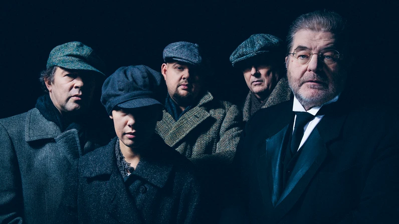 Fünf Personen in dunkler, altertümlich wirkender Kleidung und Schirmmützen stehen dicht beisammen vor schwarzem Hintergrund und blicken ernst in die Kamera, wobei Erwin Steinhauer rechts im eleganten Anzug hervorsticht.