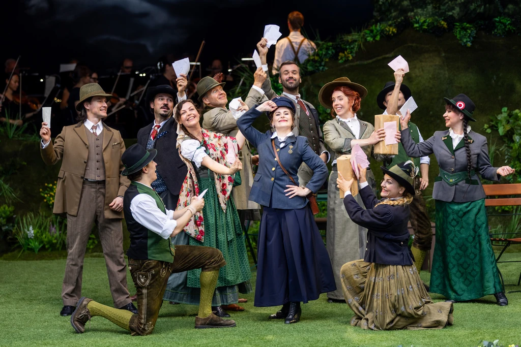 A stage photograph featuring a large group in traditional-looking costumes. The people are standing close together on a stage decorated in green. Several figures are wearing hats, waistcoats and long skirts, and are holding letters and other documents aloft whilst laughing. In the centre is a person in postal service uniform, who, whilst laughing, is also dutifully holding their right hand to their cap. In the background is a natural-looking backdrop featuring plants and a dark sky illuminated by blue lighting. The lighting highlights the entire group evenly.